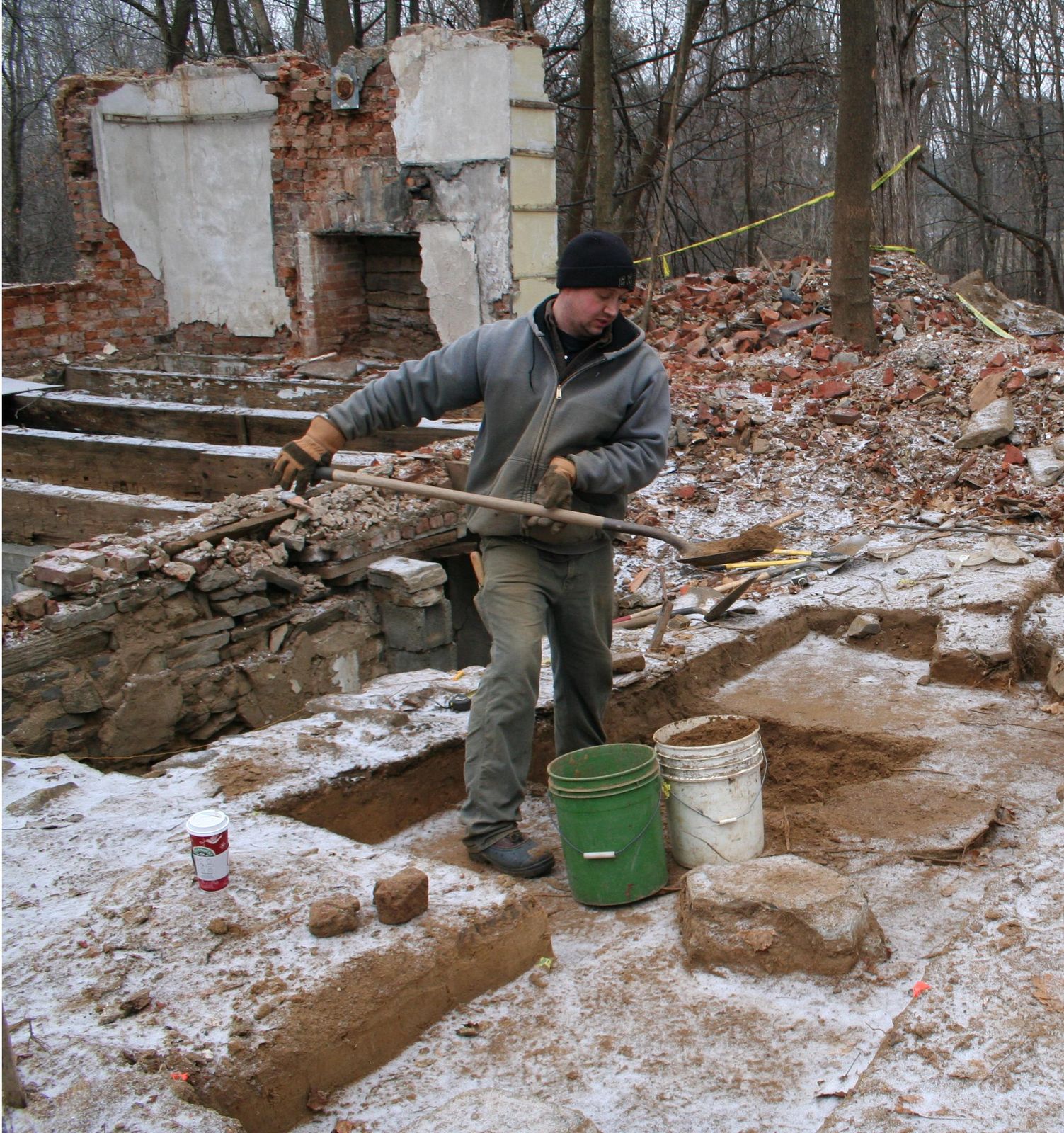 Archaeologist excavating in snow beside a stone chimney ruin